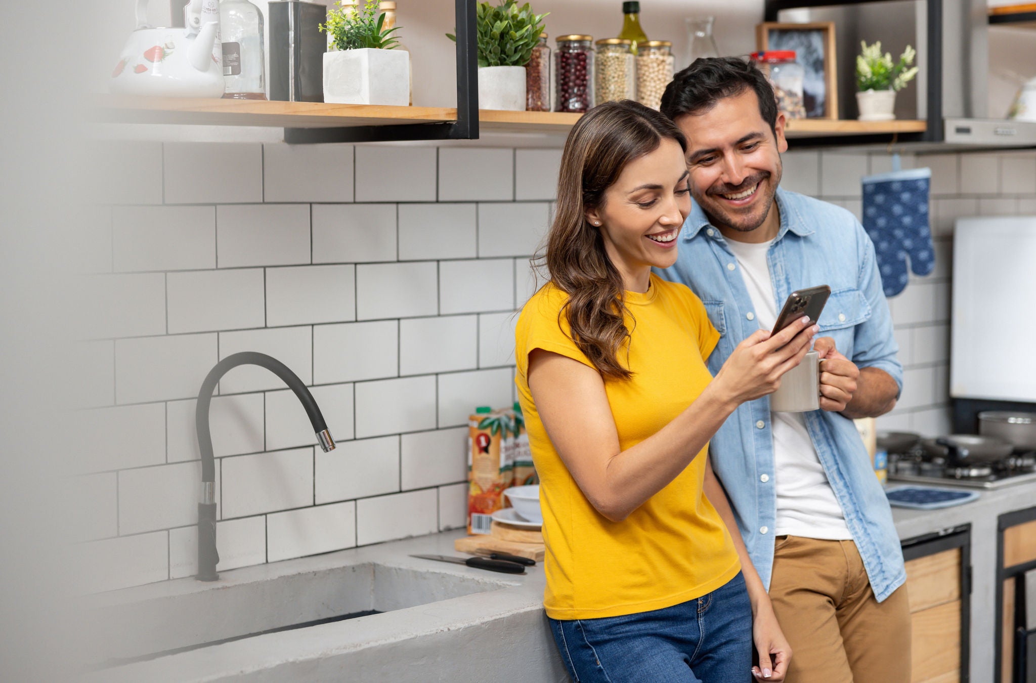 Latin American couple at home looking at social media on their cell phone while having a cup of coffee in the kitchen â   lifestyle concepts